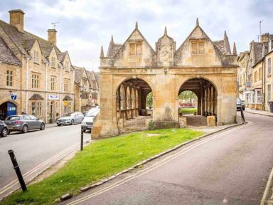 A historic stone market hall on a street with parked cars and adjacent buildings at Stanley Cottage in Chipping Campden