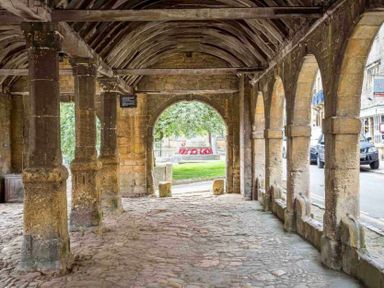 A covered stone walkway with arches and columns overlooking a street and green area at Stanley Cottage in Chipping Campden