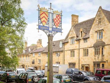 A street with historic stone buildings a wooden post with a Chipping Campden sign and parked cars