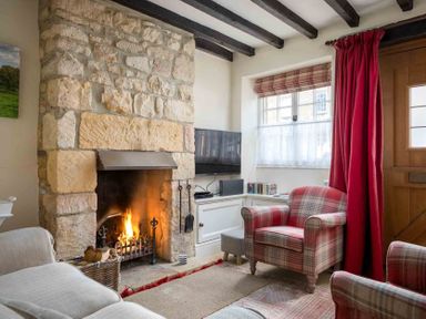 A living room with a stone fireplace a TV an armchair and red curtains at Lanes Cottage in Chipping Campden