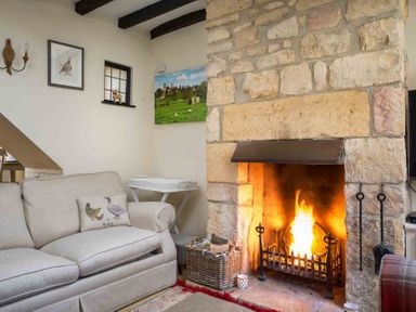 A living room with a stone fireplace with a fire a beige sofa and a small white table at Lanes Cottage in Chipping Campden