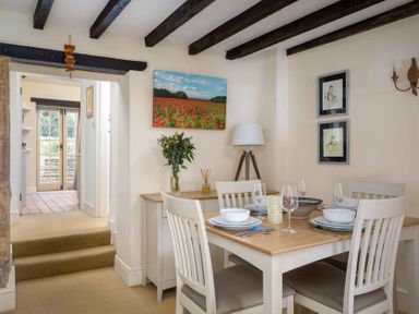 A dining room with a wooden table set for four and a sideboard with a lamp flowers and wall art at Lanes Cottage in Chipping Campden