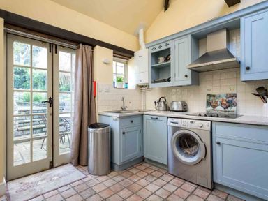 A kitchen with blue cabinets a washing machine a toaster a kettle a trash bin and glass double doors at Lanes Cottage in Chipping Campden