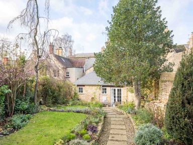 A garden path with plants and trees leading to a stone house with a slate roof and patio furniture at Lanes Cottage in Chipping Campden