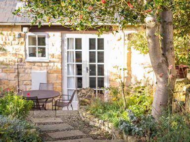 A garden with a stone pathway leading to a round table and chairs outside a stone cottage with white French doors and a window at Lanes Cottage in Chipping Campden