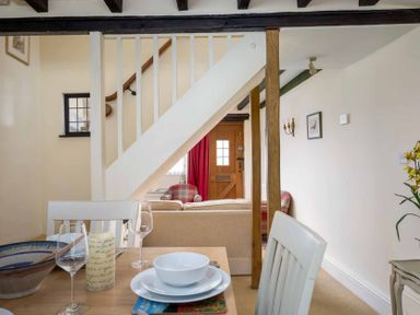 A dining area with a table set with plates and glasses next to a staircase and a living area with sofas at Lanes Cottage in Chipping Campden