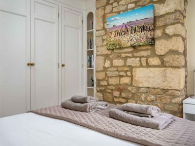 A bedroom with folded towels on a bed stone wall with a landscape picture and white wardrobes at Lanes Cottage in Chipping Campden