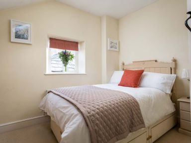 A bedroom with a bed covered with a quilt and a red cushion a window with flowers on the sill and framed pictures on the walls at Lanes Cottage in Chipping Campden