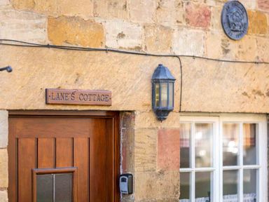 The entrance of a stone cottage with a wooden door a window a wall lamp and a sign saying Lanes Cottage at Lanes Cottage in Chipping Campden