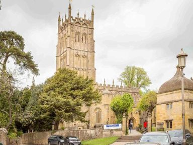 A stone church with a tall tower surrounded by trees and parked cars at Lanes Cottage in Chipping Campden