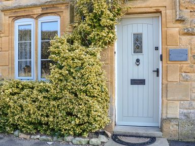A light blue front door next to tall windows with climbing plants covering the stone wall at Green Pump Cottage in Blockley