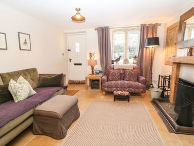 A living room with a green sofa an ottoman a purple patterned armchair a wooden side table and a fireplace at Green Pump Cottage in Blockley