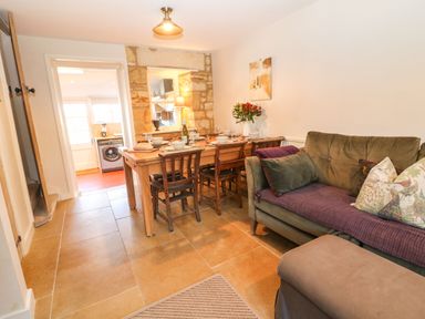 A dining area with wooden table and chairs next to a green sofa and a kitchen visible through a doorway at Green Pump Cottage in Blockley