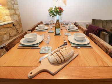 A dining table set with plates bowls glasses cutlery a bottle of wine and sliced bread on a wooden board at Green Pump Cottage in Blockley
