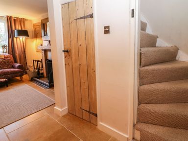 A carpeted staircase next to a wooden door and a living room with an armchair floor lamp and fireplace at Green Pump Cottage in Blockley