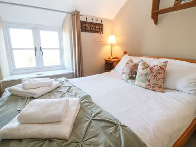 A bedroom with a wooden bed with floral pillows a bedside table with a lamp folded towels on the bed and a window in the corner at Green Pump Cottage in Blockley