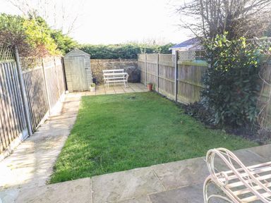 A backyard garden with a lawn wooden shed white outdoor bench and table wooden fences and paved patio at Green Pump Cottage in Blockley
