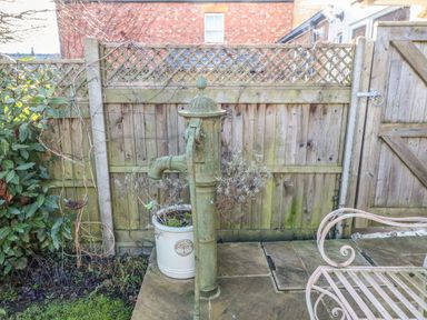 A garden area with a green metal water pump a large white plant pot with a shrub a wooden fence and a metal bench at Green Pump Cottage in Blockley