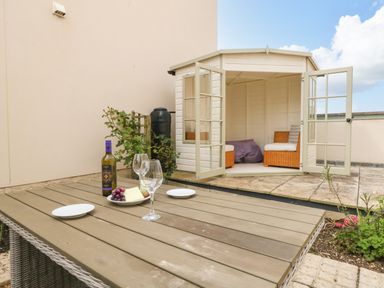 An outdoor patio with a wooden table set with wine glasses and a bottle near a white garden shed with open doors and wicker chairs inside at Pebbles Reach in Fortuneswell