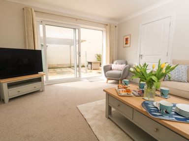 A living room with a television on a stand a coffee table with cups and flowers an armchair and a sofa near sliding glass doors at Pebbles Reach in Fortuneswell