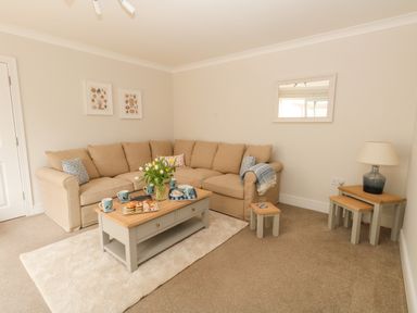 A living room with a beige corner sofa a coffee table with cups and pastries a lamp on nesting tables and wall art at Pebbles Reach in Fortuneswell
