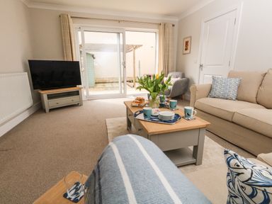 A living room with a beige sofa a gray armchair a wooden coffee table with cups and flowers and a TV on a stand at Pebbles Reach in Fortuneswell