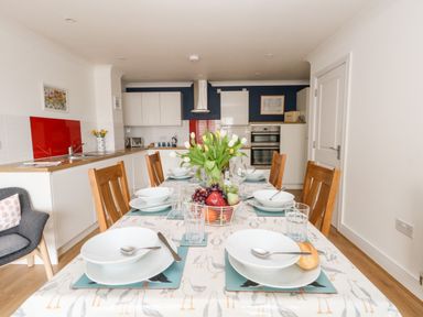 A dining table set with plates bowls glasses and cutlery in a kitchen with wooden chairs and white cabinets at Pebbles Reach in Fortuneswell