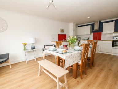 A dining area with a wooden table set for a meal and a kitchen with white cabinets in the background at Pebbles Reach Fortuneswell