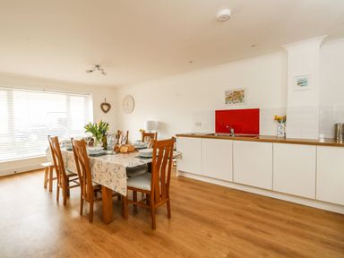 A dining area with a wooden table and chairs set with plates and glasses next to a kitchen counter with a sink and red backsplash at Pebbles Reach in Fortuneswell