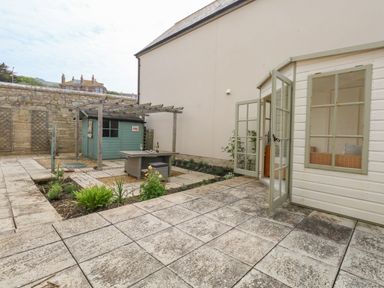 A patio with tiled flooring a white garden shed with open doors a wooden pergola a small table and chair set and a stone wall at Pebbles Reach in Fortuneswell