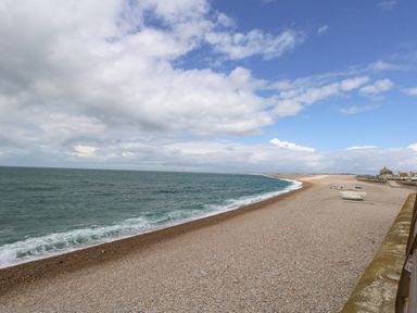A pebble beach with small boats on the shore and a sea wall on the right under a cloudy sky at Pebbles Reach in Fortuneswell