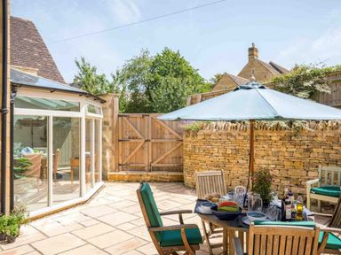A patio area with a round table set with food and drinks under a blue umbrella and wooden chairs at East Leaze in Chipping Campden