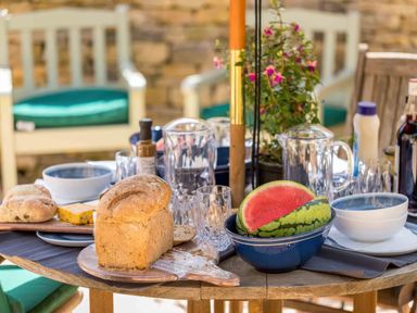 A table with bread, watermelon, glasses, bowls, bottles, and a potted plant at East Leaze in Chipping Campden
