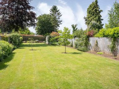 A large garden with grass lawn and small trees bordered by shrubs and wooden fences at East Leaze in Chipping Campden