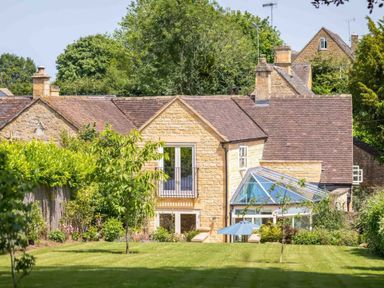 A stone house with a glass conservatory and garden with trees and shrubs at East Leaze in Chipping Campden