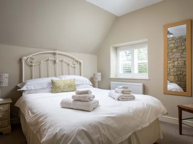 A bedroom with a white metal bed frame a bed with white linens folded towels on top a green decorative pillow two bedside tables with lamps a window with shutters a wooden framed mirror and a small wooden bench at East Leaze in Chipping Campden