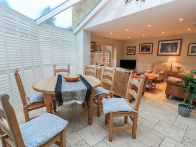 A dining area with wooden table and chairs next to a living room with sofas armchair and TV at East Leaze in Chipping Campden