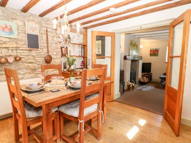 A dining area with a wooden table and chairs set for four next to a stone wall leading to a living room with a fireplace at Carters Cottage in Stow-on-the-Wold