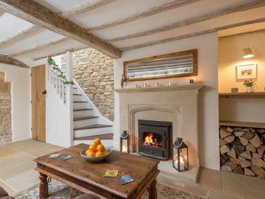 A living room with a wooden coffee table holding a bowl of oranges near a fireplace with candles and stacked firewood at Eastnor Moreton-in-Marsh