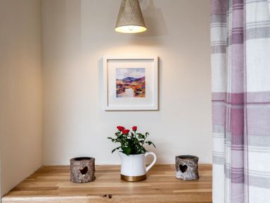 A wooden countertop with two heart cutout candle holders and a white jug with red flowers under a wall light and framed landscape painting at Eastnor in Moreton-in-Marsh