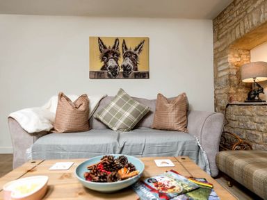 A living room with a sofa and cushions a table with magazines and a bowl of pinecones and a lamp on a stone wall Eastnor Moreton-in-Marsh