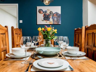 A dining room with wooden chairs a table set with plates bowls glasses and a flower vase and a cow painting on a blue wall at Eastnor in Moreton-in-Marsh