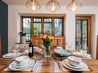 A dining table set with plates bowls glasses cutlery a wine bottle and flowers with windows and a door in the background at Eastnor Moreton-in-Marsh