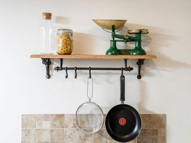 A kitchen shelf with a glass bottle corked jar of pasta and a green weighing scale with hooks below holding a strainer and a frying pan at Eastnor in Moreton-in-Marsh
