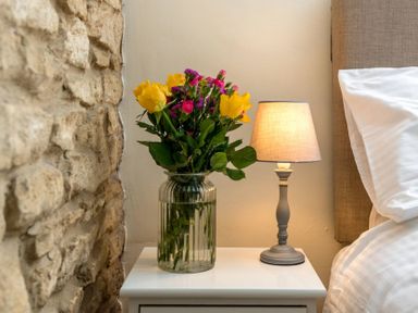 A bedside table with a flower vase and a lit lamp next to a bed at Eastnor in Moreton-in-Marsh