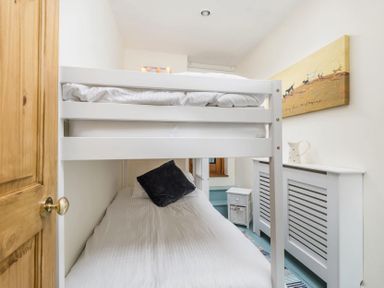 A bedroom with a white bunk bed a black pillow a small white side table a radiator cover and a wall painting at Eastnor in Moreton-in-Marsh