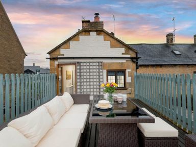 An outdoor seating area with cushioned wicker furniture a glass table and wooden picket fences at Eastnor in Moreton-in-Marsh