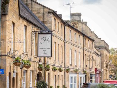 Stone building with multiple windows and hanging flower baskets on a street with cars at Eastnor in Moreton-in-Marsh