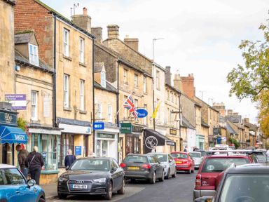 A street with parked cars and stone buildings with various shops and people walking at Eastnor in Moreton-in-Marsh