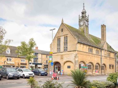 A street view with parked cars trees and a historic clock tower building at Eastnor in Moreton-in-Marsh